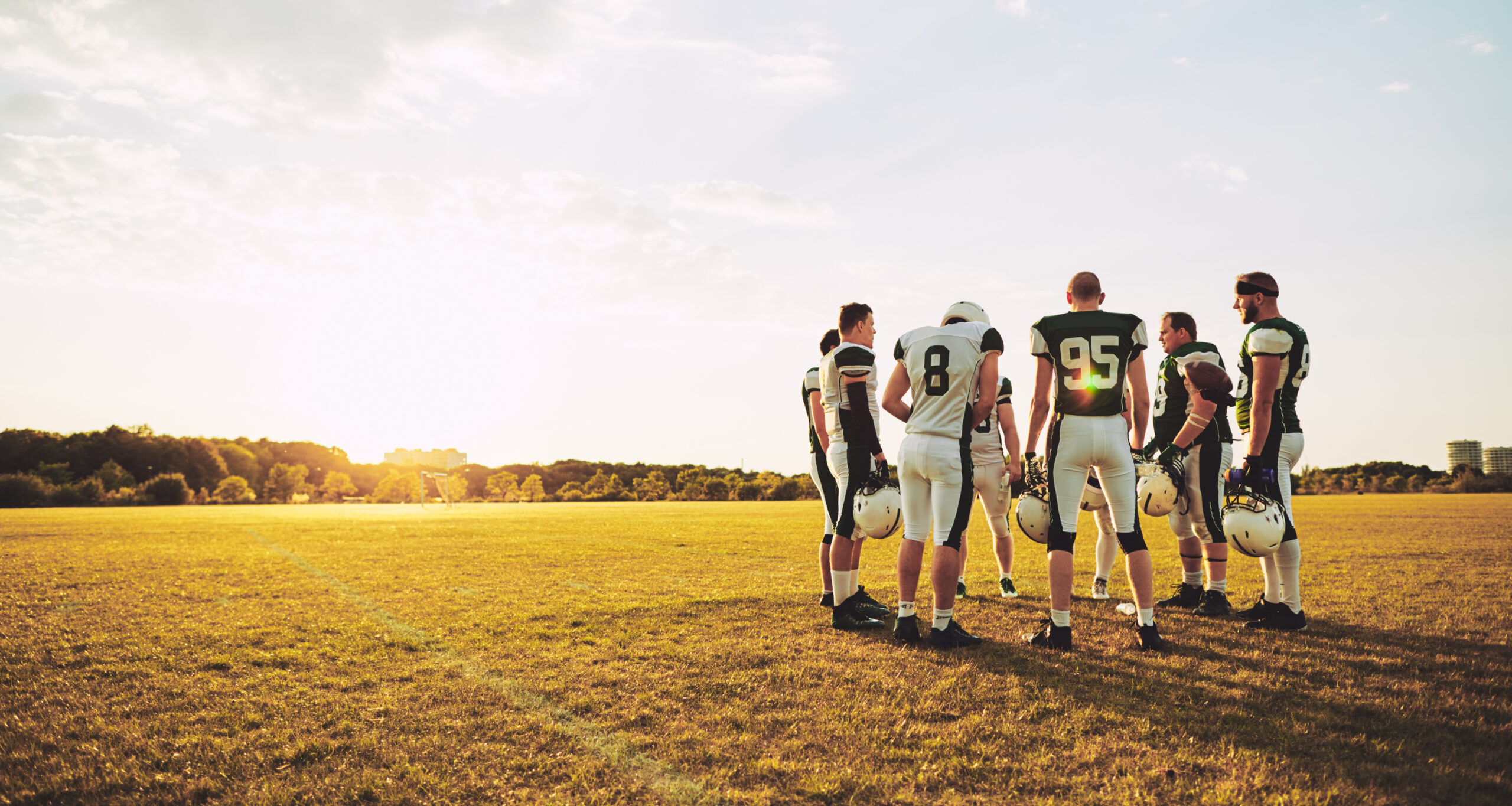 A football team huddled up on a field with the sun shinning behind them.